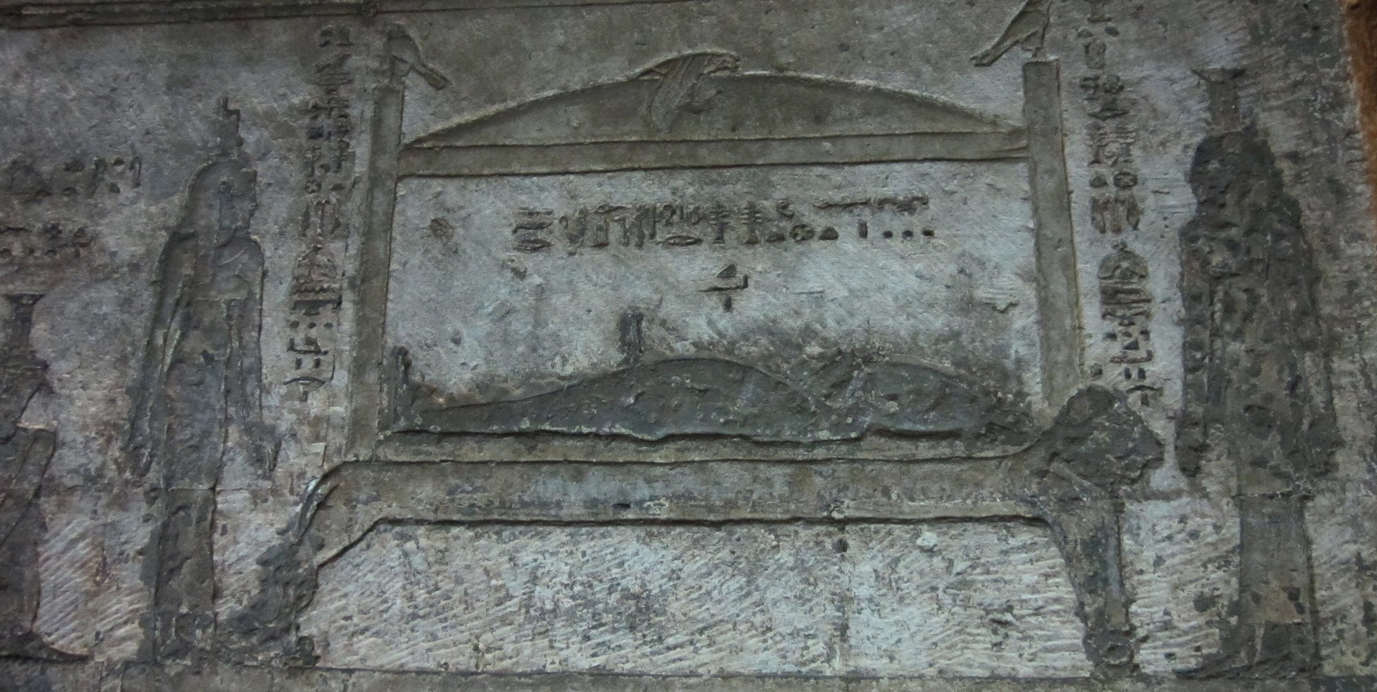 An ancient Egyptian sunk-relief scene carved in stone: the dead god Osiris lies wrapped as a mummy on a low funerary bier beneath an arched shrine-canopy, a bird (the soul as a hawk) hovering with outspread wings over the canopy's ridge; a tall crowned goddess stands mourning at the left and another at the right, each with columns of hieroglyphs above, the whole panel worn grey temple limestone.