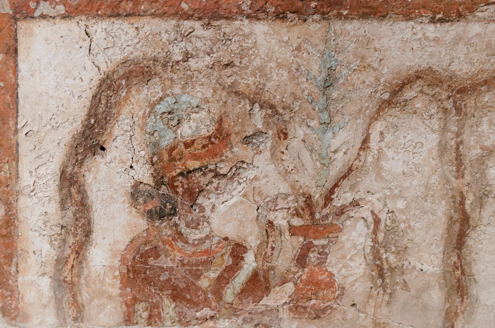 Detail of the Mari palace wall painting known as the Investiture of Zimri-Lim: a goddess in a blue-crowned headdress and a red-and-blue flounced robe stands within a painted red frame, holding a globular vessel from which a leafy plant and streams of water rise — the flowing-vase goddess who embodies the life-giving sweet waters, the abundance of the water-god Ea. The surface is a cracked, fragmentary tempera-on-plaster ground in ochre, red and turquoise.