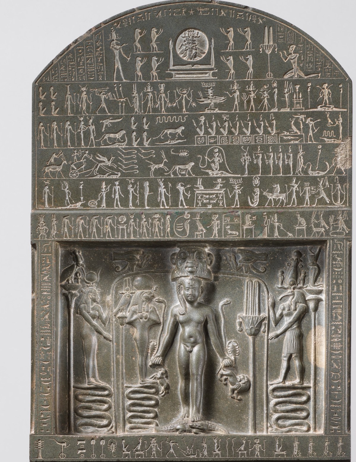 Ancient Egyptian dark-stone magical stela (cippus): in a recessed panel the nude child-god Horus stands frontally on two crocodiles, grasping serpents, a scorpion, a lion and a gazelle in his fists, crowned by the mask of the god Bes and flanked by two deities standing on coiled serpents; above, an arched lunette with registers of gods and dense hieroglyphs.