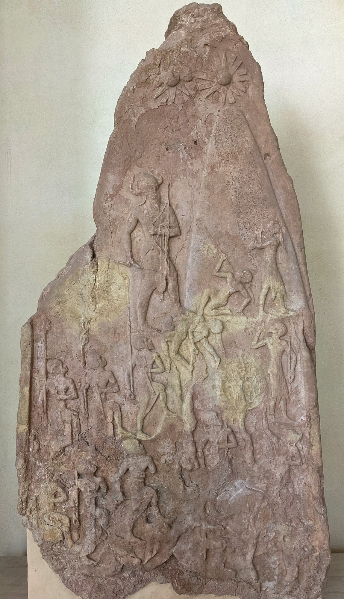 The Victory Stele of Naram-Sin: a tall pink-sandstone monument carved in relief. The Akkadian king Naram-Suen, wearing a horned helmet of divinity and carrying a bow and spear, climbs a wooded mountain above his soldiers, who advance in file; his defeated enemies fall, flee and beg beneath him, and sun-and-star glyphs of the gods shine over the peak.