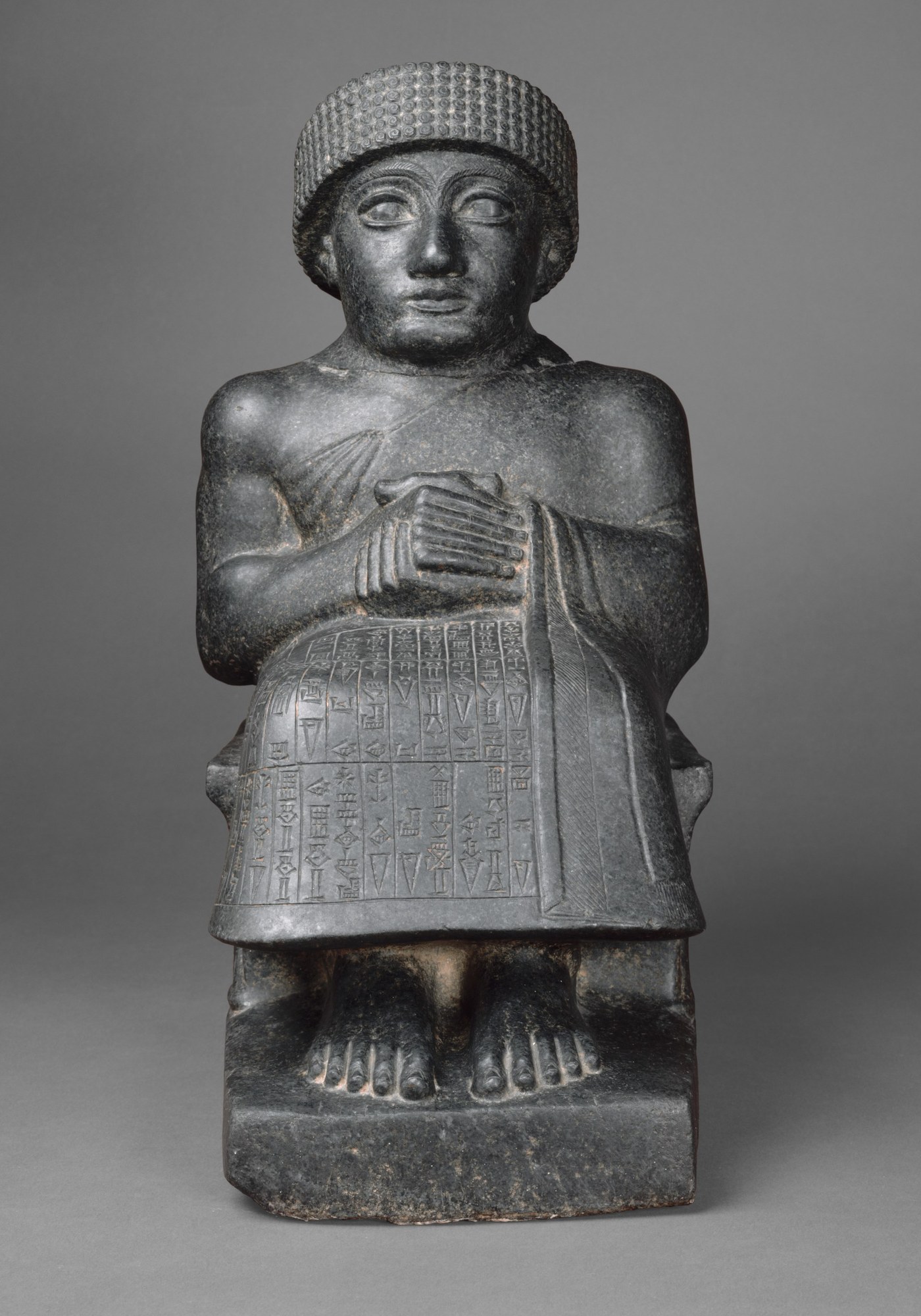 Neo-Sumerian diorite statue of the ruler Gudea of Lagash, seated with hands clasped in prayer at his chest, wearing a brimmed fleece cap and a fringed cloak over one shoulder; his lap and skirt are covered with columns of cuneiform, his bare feet resting on the base.