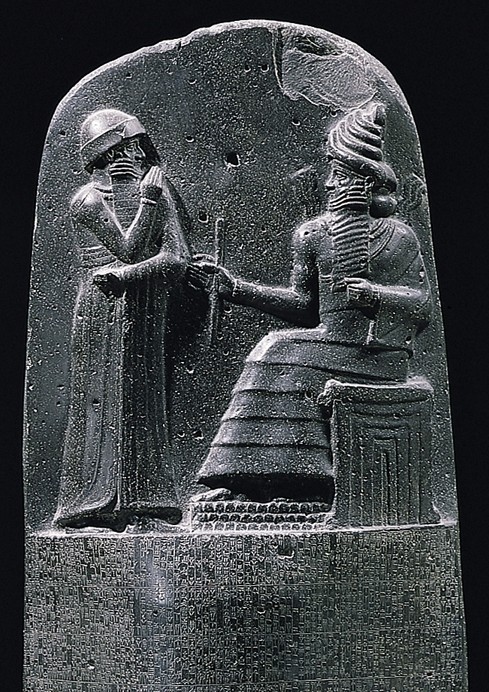 The relief at the top of the law stele of Hammurabi: at right the sun-god Shamash sits enthroned on a tiered temple-façade throne, wearing the many-tiered horned crown of divinity with flames rising from his shoulders, extending the rod-and-ring of just rule; at left the standing king Hammurabi raises one hand to his mouth in reverence to receive it. Carved in dark basalt, the figures set against a plain black ground above the opening lines of the cuneiform law-code.
