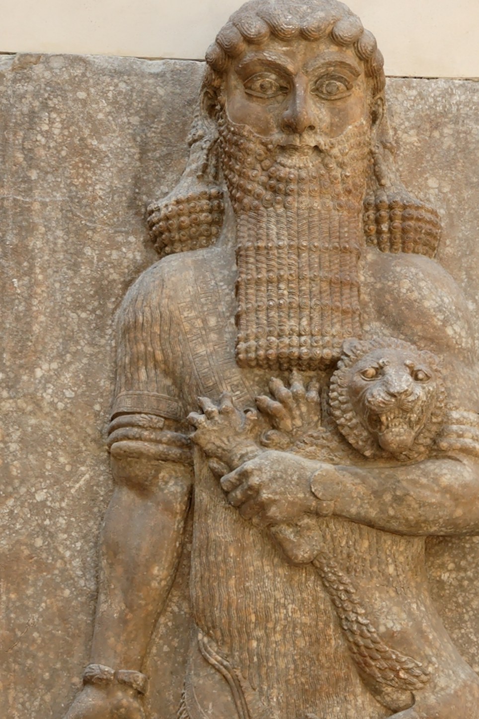 Neo-Assyrian stone relief: a colossal bearded male figure with elaborately curled hair and a long tiered beard stands frontally, cradling a small lion against his chest with his left arm while a coiled object (read as a serpent) hangs from his lowered right hand; carved gypsum-alabaster from a palace façade.
