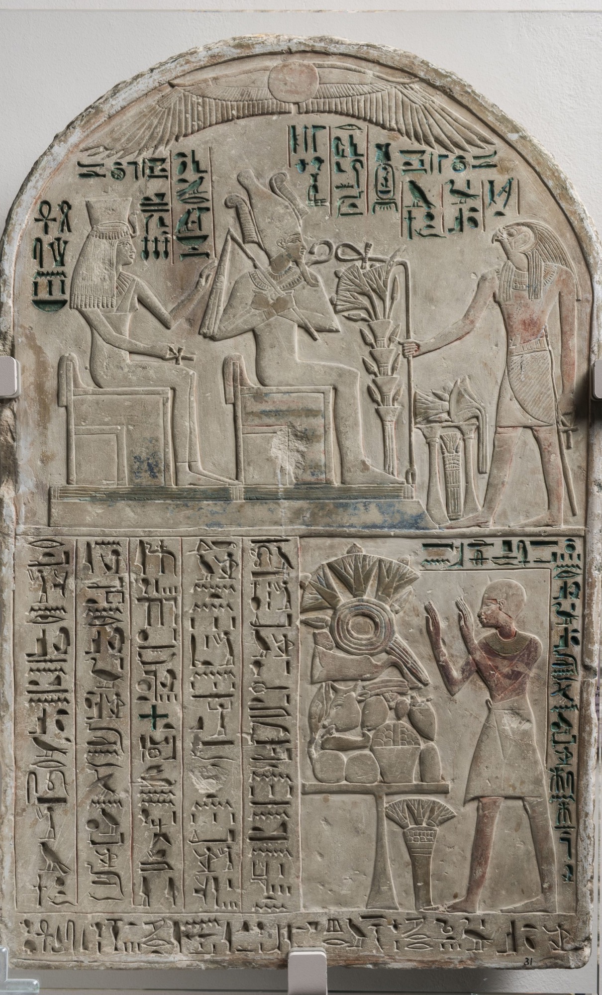 Ancient Egyptian limestone stela with an arched, painted lunette. Under a winged sun-disk, the upper register shows the god Osiris enthroned in the tall White Crown holding the crook, the goddess Isis seated behind him on a second throne, and the falcon-headed god Horus standing before them presenting lotus flowers over a laden offering-stand; columns of green-filled hieroglyphs fill the field, with a royal cartouche between Osiris and Horus. In the lower register a shaven-headed worshipper raises both hands in adoration before a heaped offering-table, beside several columns of a hieroglyphic hymn.