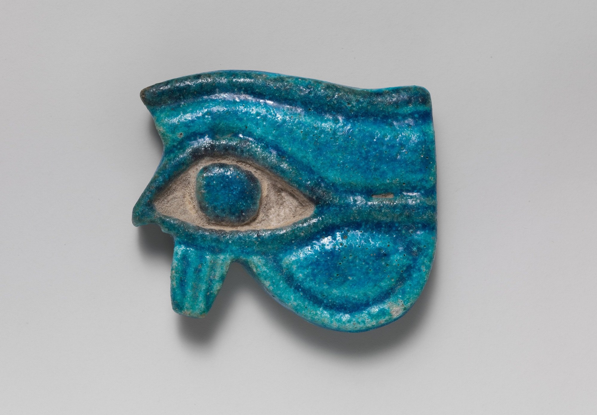 An ancient Egyptian wedjat (Eye of Horus) amulet in brilliant turquoise-blue glazed faience, photographed on a plain neutral ground. The stylised falcon-eye shows the arched brow and dark upper cosmetic-line, the almond eye with a raised round pupil, a vertical striated teardrop marking dropping straight down below the eye, and a curling spiral cheek-marking sweeping out to the right. The glaze is worn to pale matte around the eye socket; the whole amulet is intact.