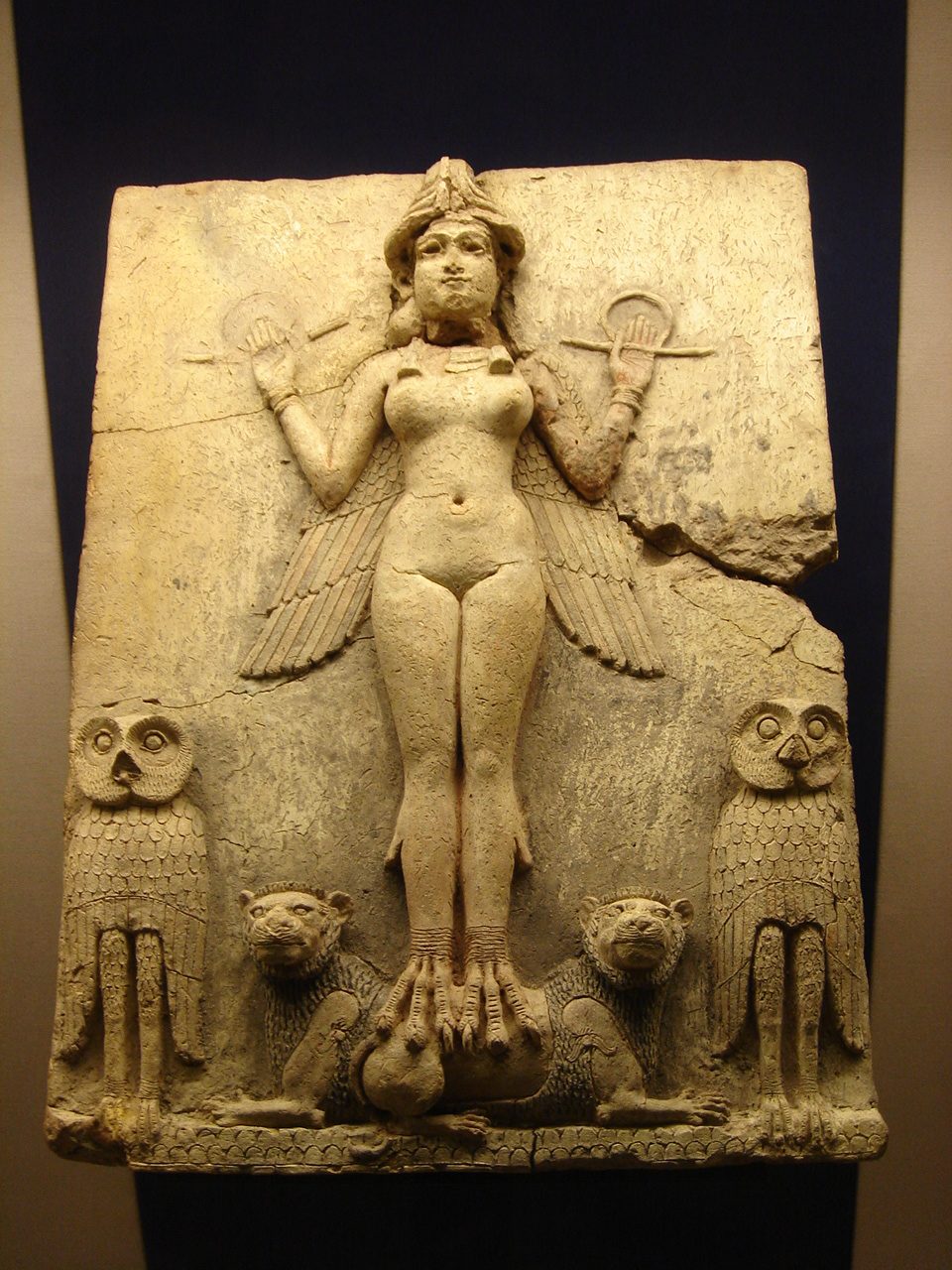Old Babylonian terracotta relief plaque: a frontal nude winged goddess with a horned crown and bird talons for feet, holding a rod-and-ring measuring emblem in each raised hand, standing on the backs of two recumbent lions and flanked by a pair of upright owls, over a scale-patterned mountain register.