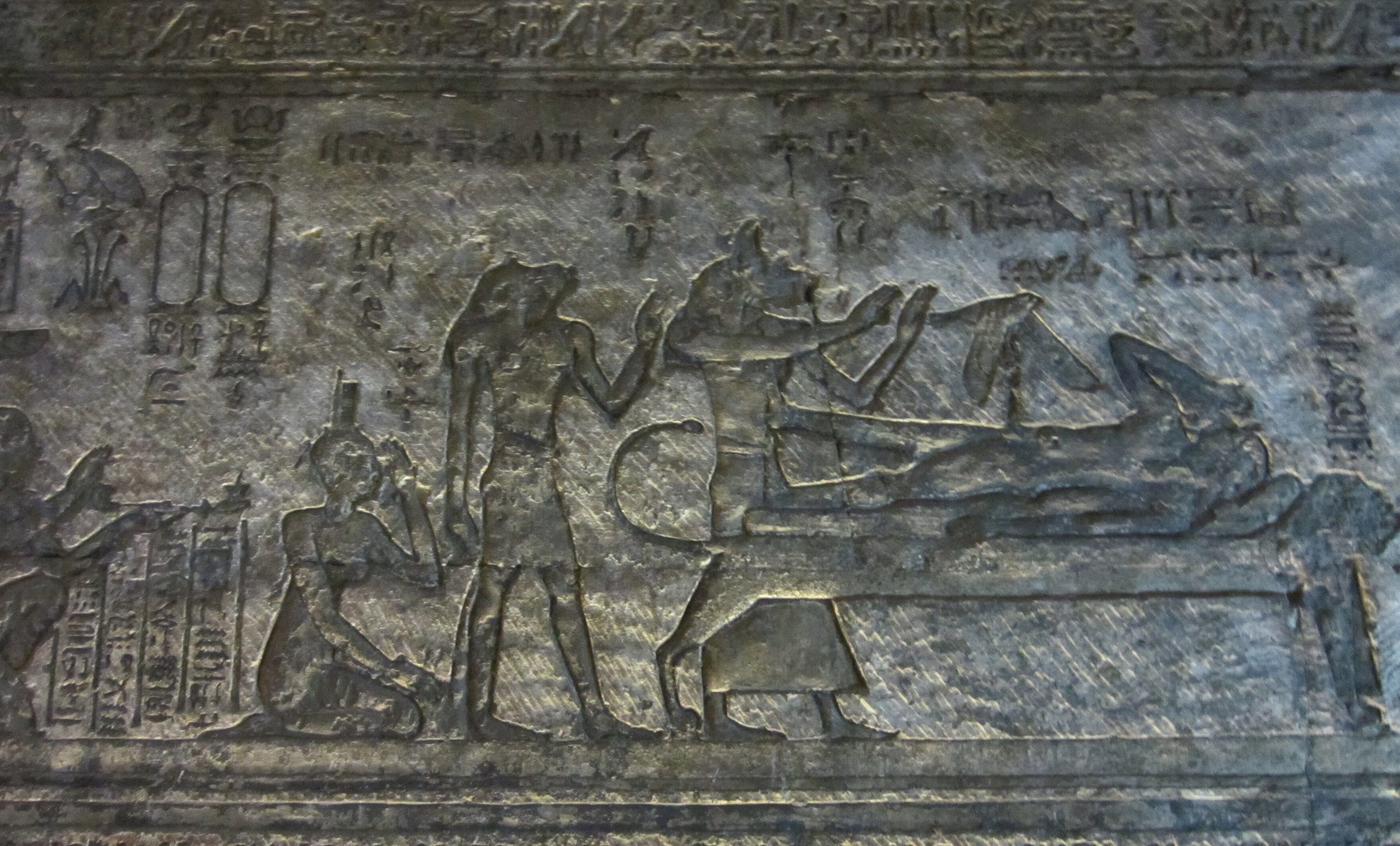 Ancient Egyptian temple relief carved in raised relief on the chapel wall. At right the god Osiris lies recumbent on a lion-couch bier; the jackal-headed Anubis bends over the body to revive it, while behind him a standing goddess raises a hand and a kneeling goddess mourns at the couch's foot. Columns of hieroglyphs and royal cartouches fill the field above, framed by a hieroglyphic border band.