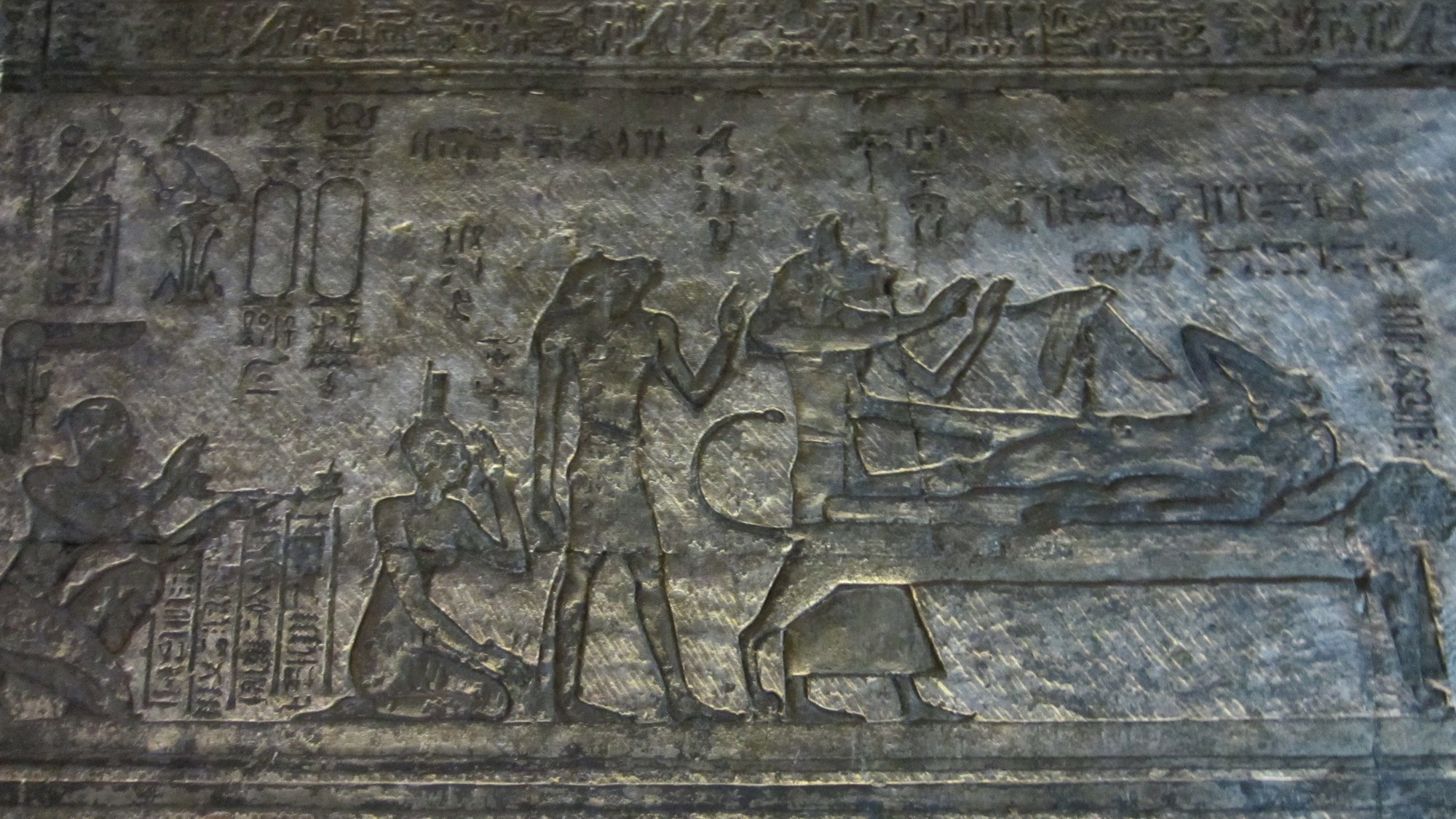 Ancient Egyptian temple relief carved in sunk relief on a chapel wall. At right the god Osiris lies recumbent and ithyphallic on a low bier; above his body the goddess Isis, in the form of a kite with outstretched wings, settles to conceive the heir. The jackal-headed god Anubis attends at the couch, and to the left a standing goddess and a kneeling goddess mourn, with a seated figure at the far edge. Columns of hieroglyphs and royal cartouches fill the field, framed by hieroglyphic border bands above and below.