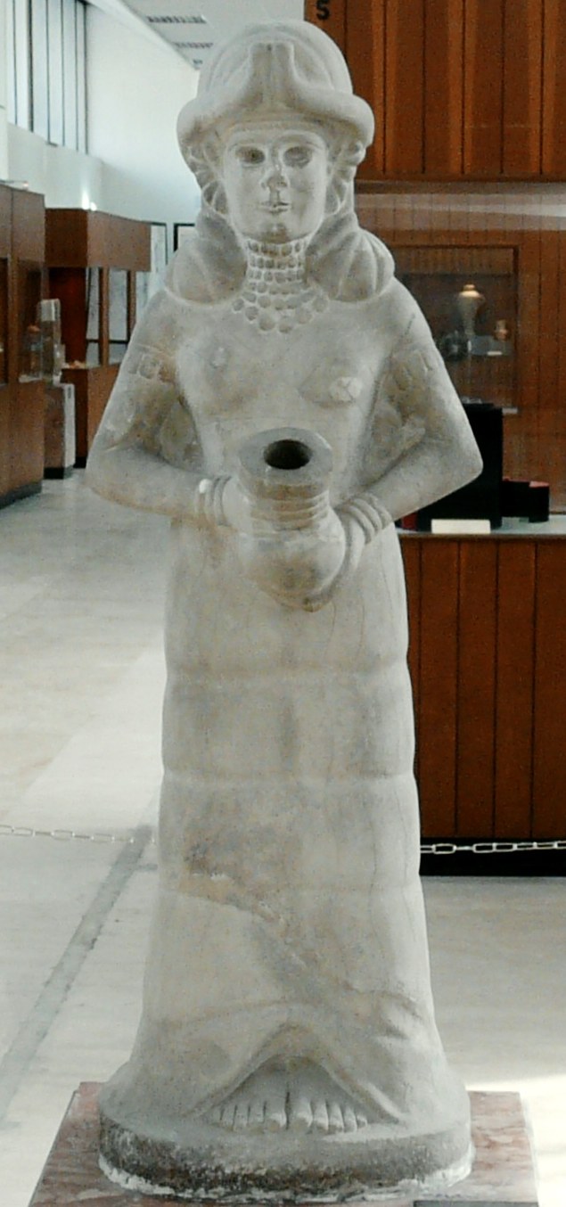 Weathered ancient limestone statue of a standing goddess in a horned headdress and a long flounced robe, a tiered bead choker and necklace at her throat, holding at her belly in both hands a round vase pierced through its centre — the vessel from which the life-giving waters were made to flow; displayed on a plinth in the National Museum of Aleppo.
