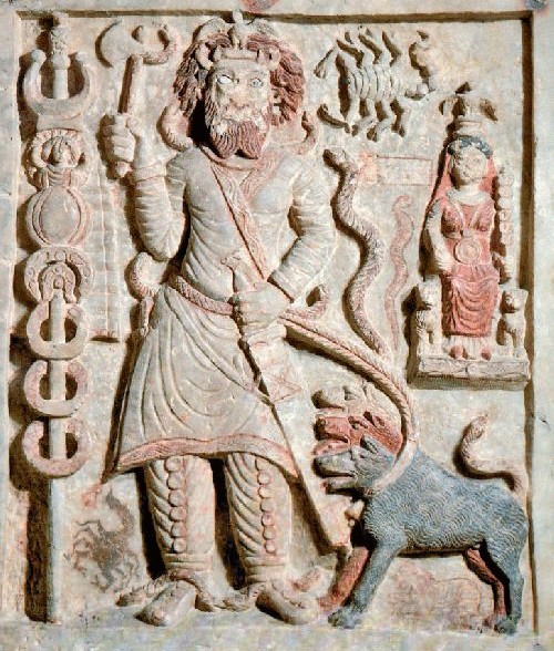 Carved stone relief with traces of red pigment: a bearded male god in a spiked crown stands frontally, raising a mace in his right hand with a dagger at his belt, flanked by tall standards; a large hound sits at his feet and a scorpion, a coiled serpent and a small enthroned attendant appear to the right — the Mesopotamian god Nergal with his underworld emblems.