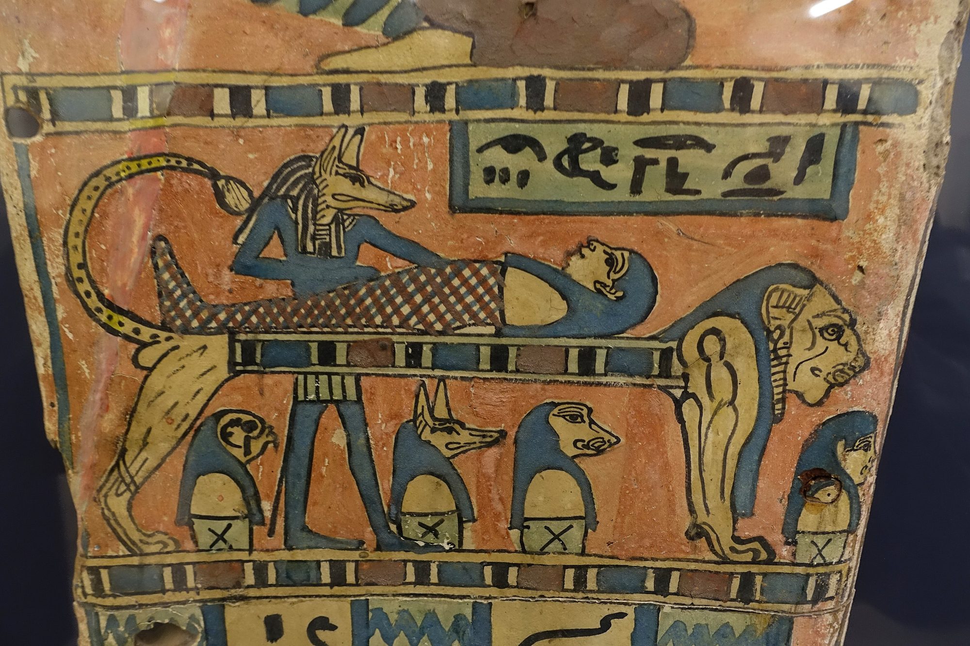 An ancient Egyptian painted coffin fragment: the sky-goddess Nut spreads her feathered wings above the mummy tended by jackal-headed Anubis, hieroglyphs around them.