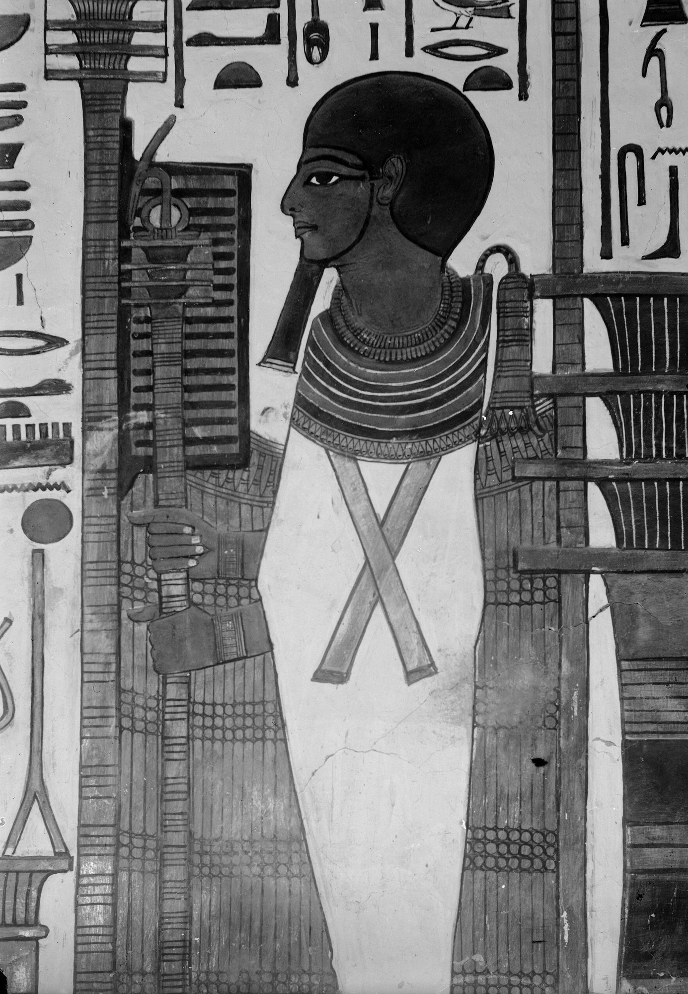 Wall-painting of the god Ptah in his shrine: a mummiform figure in a close skullcap and straight divine beard, a broad beaded usekh collar over his shoulders, his hands emerging from the wrapped body to grip a tall composite was-djed-ankh staff, standing within a kiosk framed by djed-pillars and columns of hieroglyphs. Black-and-white archival photograph of the ancient painting.