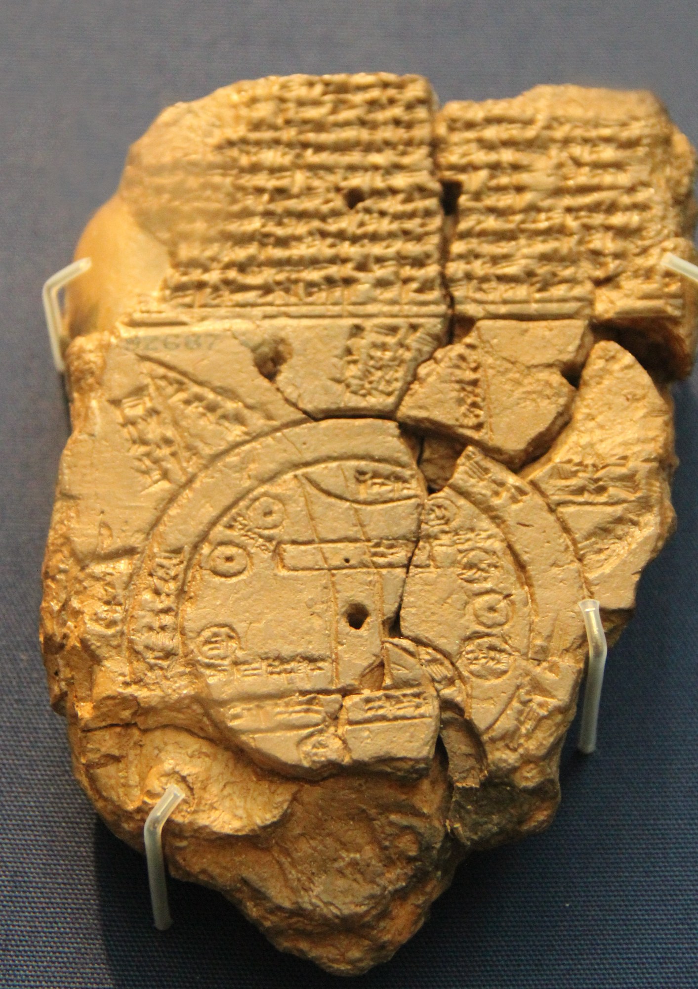 A broken Babylonian clay tablet, its upper half filled with lines of cuneiform and its lower half incised with a circular map of the world: the earth as a disc ringed by the ocean, with radiating triangular regions beyond it.