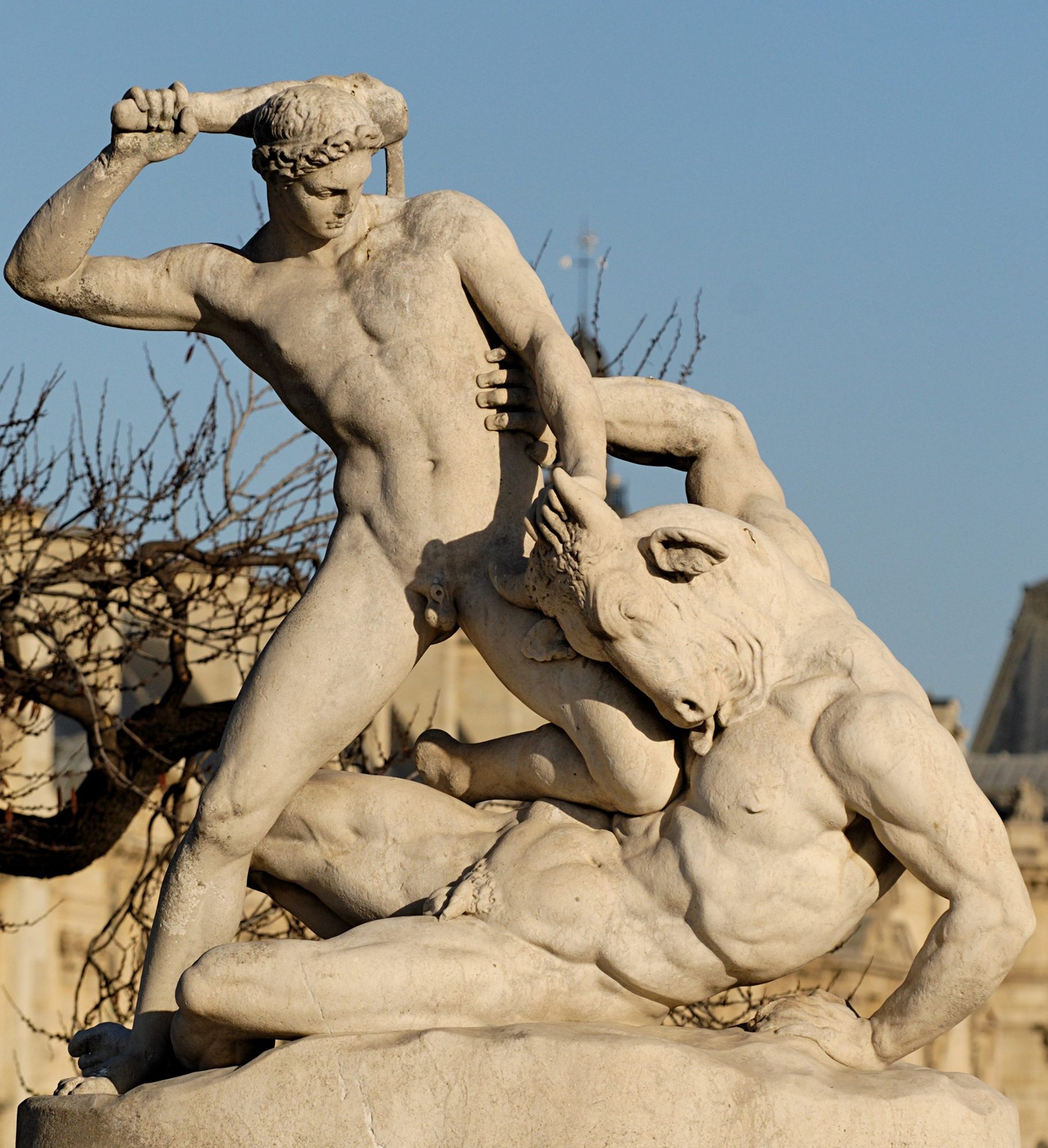 A white marble statue of a nude, laurel-crowned Theseus lunging forward with a club raised overhead in one hand, gripping the horned head of the bull-headed Minotaur with the other as the beast collapses beneath him over a fallen youth, set against a soft-focus ground of bare trees and pale stonework.