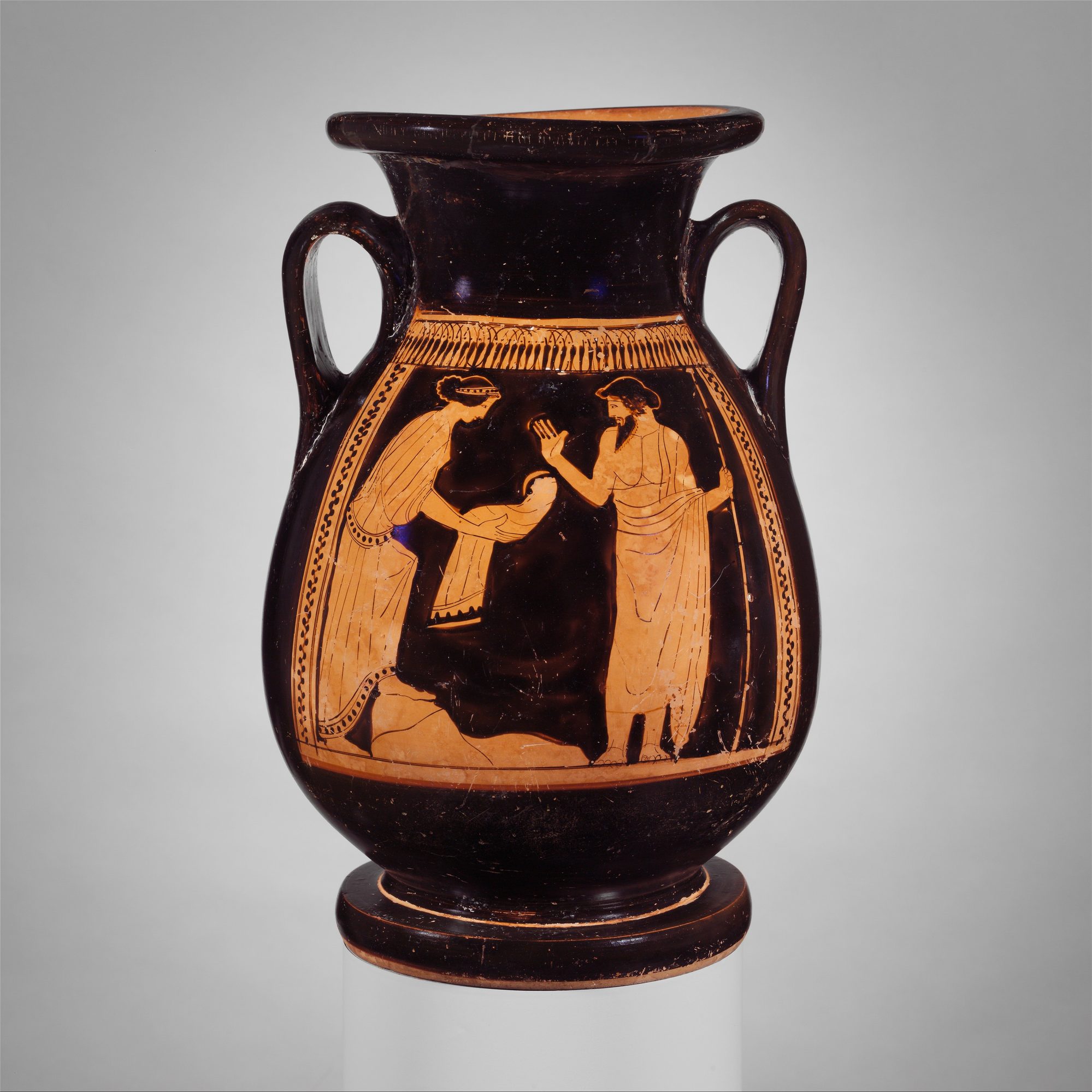 An Attic red-figure pelike, the whole black-glazed vessel standing against a plain grey ground, its belly panel bordered by a band of tongues above and running dot-chains at the sides. In the scene the Titaness Rhea, at left in a long pleated chiton and mantle with her hair bound in a fillet, steps forward and holds out in both hands the stone wrapped in swaddling clothes; facing her at right the bearded Kronos, in a himation that leaves one shoulder bare and a soft cap, grips a long sceptre in his trailing hand and raises his other hand to receive the bundle he believes is his newborn son — the swaddled-stone deception that founds the ruling family.
